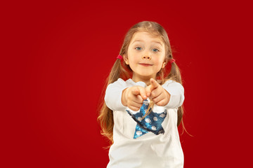 Beautiful emotional little girl isolated on red background. Half-lenght portrait of happy child showing a gesture and pointing up. Concept of facial expression, human emotions, childhood.