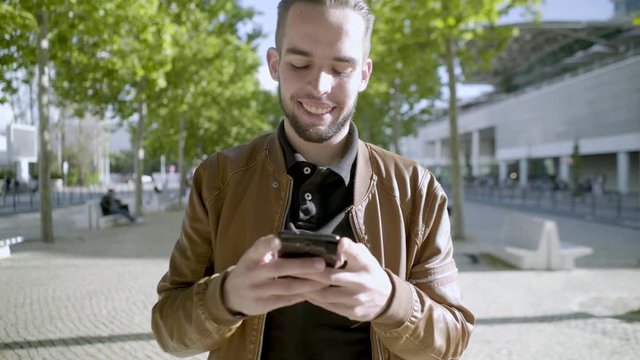 Smiling young bearded man using smartphone during stroll. Cheerful brunet looking at phone while walking on park alley. Technology concept