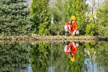 Fototapeta na wymiar Take care of nature and save planet for future generations. Little girl in red raincoat and gas mask is squatting on bank of pond. Evergreen trees and girl are reflected in mirror of pond.