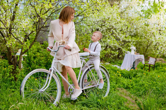 young mother of son sit together playing actively having fun on white old bike in blooming garden spring flowers during summer holidays.