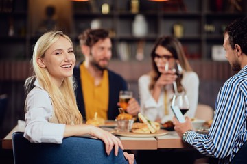 Young smiling Caucasian blonde cute woman dressed smart casual sitting at table in restaurant and looking over shoulder. In background are her friends having dinner.