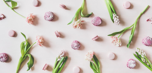 Flat-lay of sweet pink macaron cookies, lilac marshmallows and fresh spring flowers over pastel pink background, top view. Food texture, background and wallpaper