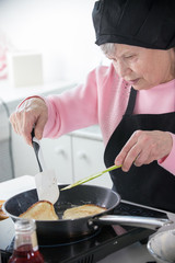 An old woman in a cap and apron frying pancakes in the kitchen. Turn over the pancakes