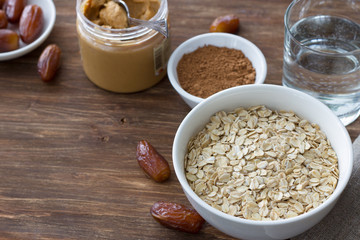Raw oatmeal in a white bowl, dates fruits, peanut butter, cocoa and a glass of water, ingredients for delicious healthy breakfast