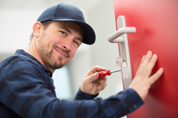 happy young carpenter installs door lock with wireless screwdriver - obrazy, fototapety, plakaty