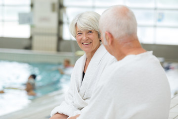 elderly couple wearing bathrobes sat by indoor pool
