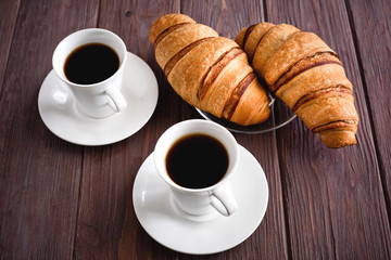 Two croissants on a saucer and two black coffees on a brown wooden table. 