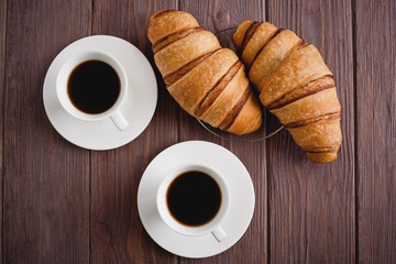Breakfast for two. Two croissants on a saucer and two black coffees on a brown wooden table. Top view