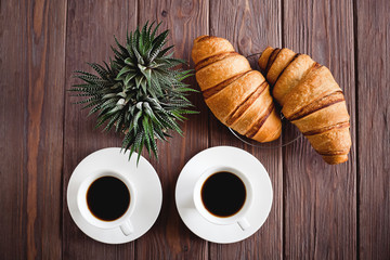 Two croissants on a saucer and two black coffee in white cups on a brown wooden table. Cactus in a pot. Breakfast. View from above