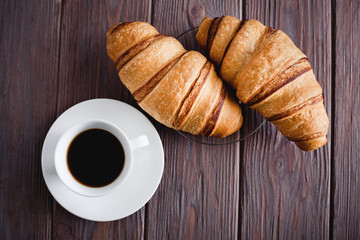 Breakfast. Two croissants on a saucer and black coffee in a white cup on a brown wooden table. View from above.
