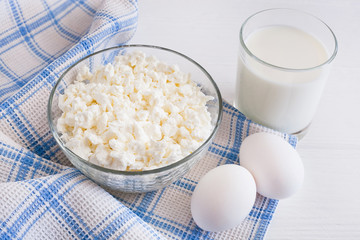 Fresh cottage cheese in a bowl with a glass of milk and eggs on a white wooden table, blue towel in a cage. top copy space