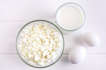 Fresh cottage cheese in a transparent bowl with eggs and a glass of milk on a white wooden background, top view