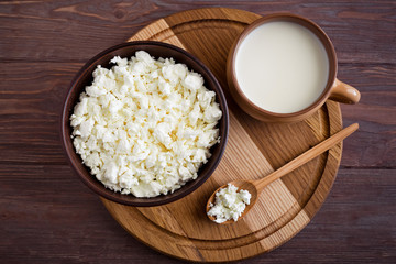 bowl with cottage cheese and spoon with cottage cheese lies on a wooden board with a cup of milk on a brown table, top view