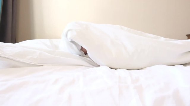 funny little boy peeks out from under soft blanket smiles and covers blanket with head in large parents bed slow motion