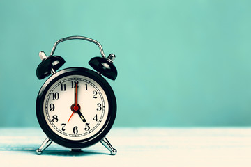 Retro black alarm clock on turquoise background with copy space. Alarm at 7 o'clock clock hand at 5...