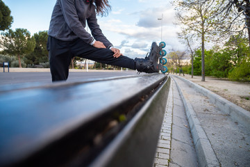 Fototapeta premium From below side view of young cool man in sportswear with roller skates stretching on bench in park