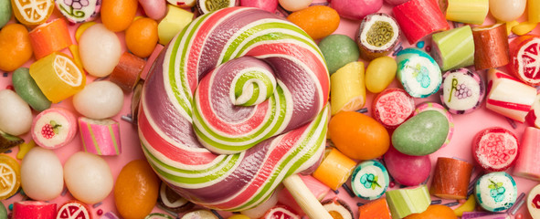 Panoramic shot of multicolored round lollipop on wooden stick near fruit caramel candies on pink...