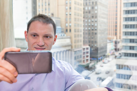 Closeup of young happy man businessman sitting at window in home room taking selfie with phone smiling in NYC