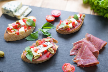 Traditional Italian bruschetta with blue cheese, feta, tomatoes, basil leaves, jamon on black stone background.