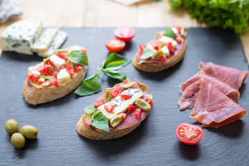 Traditional Italian bruschetta with blue cheese, feta, tomatoes, basil leaves, jamon on black stone background.