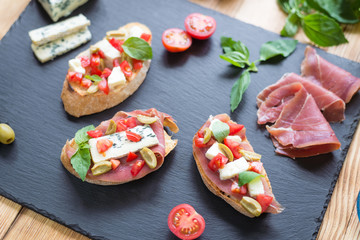 Traditional Italian bruschetta with blue cheese, feta, tomatoes, basil leaves, jamon on black stone background.
