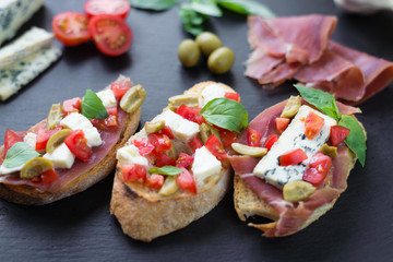 Traditional Italian bruschetta with blue cheese, feta, tomatoes, basil leaves, jamon on black stone background.