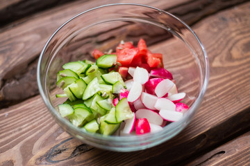 Sliced fresh cucumbers, tomatoes, red radishes and seeds in a glass plate on a wooden background. Cooking salad from vegetables. The concept of healthy eating, vegetarianism.