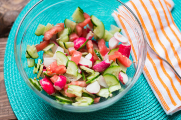 Cooking salad from fresh vegetables. Sliced fresh cucumbers, tomatoes, red radishes and seeds in a glass plate on a turquoise background. The concept of healthy eating, vegetarianism.