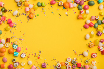 frame of multicolored candies and sprinkles scattered on yellow background with copy space