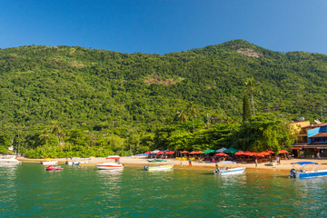 Fototapeta na wymiar Village by the sea with a tourist beach, people on the sand, red umbrellas and the warm crystal sea water with tropical jungle behind with taxi boats on a summer sunny day, on an island in Brazil
