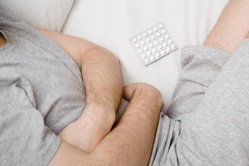 Man lying down on his side in bed. Hands touching belly and holding pack of white pills. Suffering from abdominal pain at home. Stomach ache problems and solution concept. Close up. Top view. 