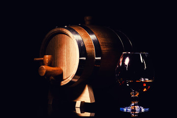 Glass of cognac with oak barrel on black background. Wooden barrel for whiskey with glass.