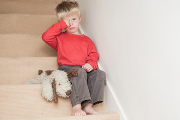 Lonely upset child sitting on the stairs in the home with his teddy next to him.