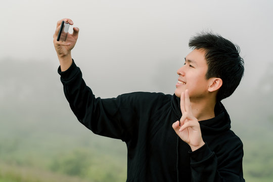 Asian boy teenager wearing black winter clothing Stand to take pictures of yourself with a mobile phone On the road along the reservoir in the morning mist And morning sunlight Happy expression
