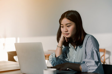 Asian woman Wearing a blue striped shirt sitting in front of a laptop computer Take her hands, feet, chin With the expression being thinking about work