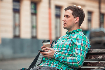 portrait of young handsome man sitting on bench drinking coffee browsing internet via phone