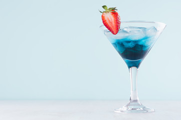 Fresh exotic alcohol drink with blue liquor curacao, strawberry, ice cubes in wineglass on elegant blue pastel color background.