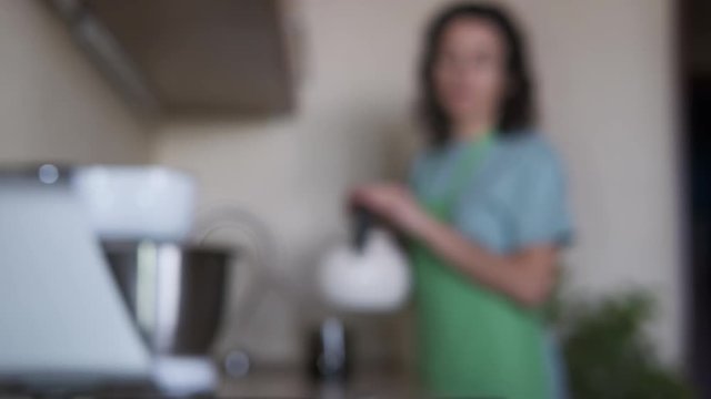 A woman in the kitchen brewing tea. A woman is putting a hot kettle in front of the camera.