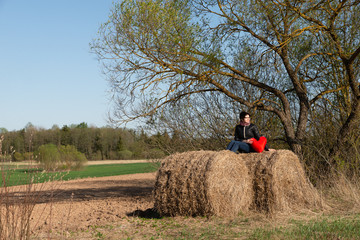 Lonely whomen is siting on the hay. On the background of the countryside near a tree. Solitude. Holding a red heart.