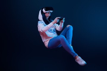 young woman in virtual reality headset levitating and holding joystick on blue - obrazy, fototapety, plakaty