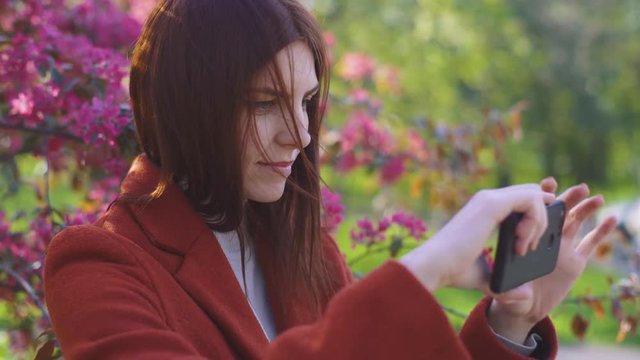 Young attractive red-haired woman makes a selfie on the background of spring flowers of cherry or sakura blossoms on smartphone at sunset in park 