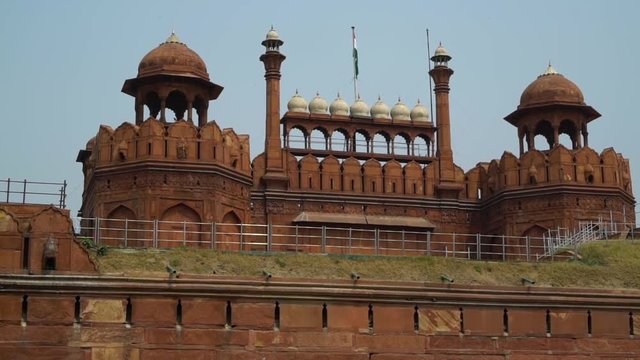 At the Red Fort (Lal Qila) during the day.  Indian National flag waves is on a flag pole off the  roof of the fort. Handheld. Wide shot. (WS).