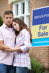 Young Couple Forced To Sell Home Through Financial Problems Standing Outdoors Next To For Sale Sign