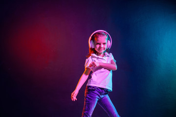 Girl of 7 years old listening to music in headphones and jump on dark colorful background. Neon light. Dancing girl. Happy small girl dancing to music. Cute child enjoying happy dance music.