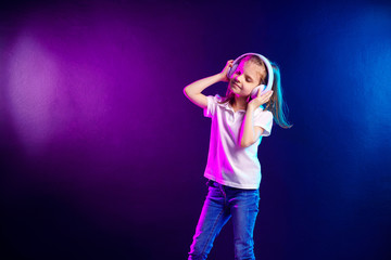 Girl listening to music in headphones on dark colorful background. Cute child enjoying happy dance music, close eye's and smile posing on studio background wall.