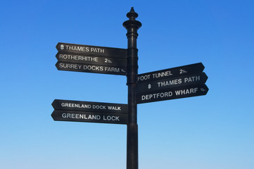 Black iron signpost giving directions to the Thames Path, Rotherhithe, Surrey Docks Farm, Greenland Dock Walk, Greenland Lock, Foot Tunnel, Thames Path, and Deptford Wharf in East London.