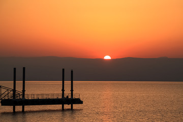 Sun peeks over the mountains along the Sea of Galilee as tourist sits on pier to watch