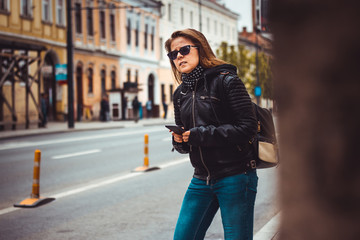 Plakat Brunette young woman wearing jeans, leather jacket and sunglasses – Girl with backpack hailing for a car or using phone to find directions and guidance during travel – Concept image for car sharing