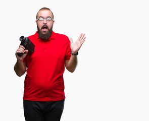 Young caucasian hipster man filmming using retro video camera over isolated background very happy and excited, winner expression celebrating victory screaming with big smile and raised hands