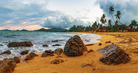Cloudy evening view of Maenam beach, Koh Samui, Thailand. Beautiful seascape with sea waves and rocks scattered on beach sand. Rainy ominous grey storm clouds - dramatic sky.